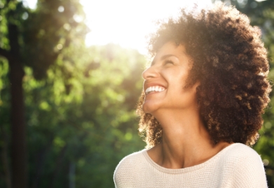 Woman grinning outdoors with sunlight streaming through trees behind her